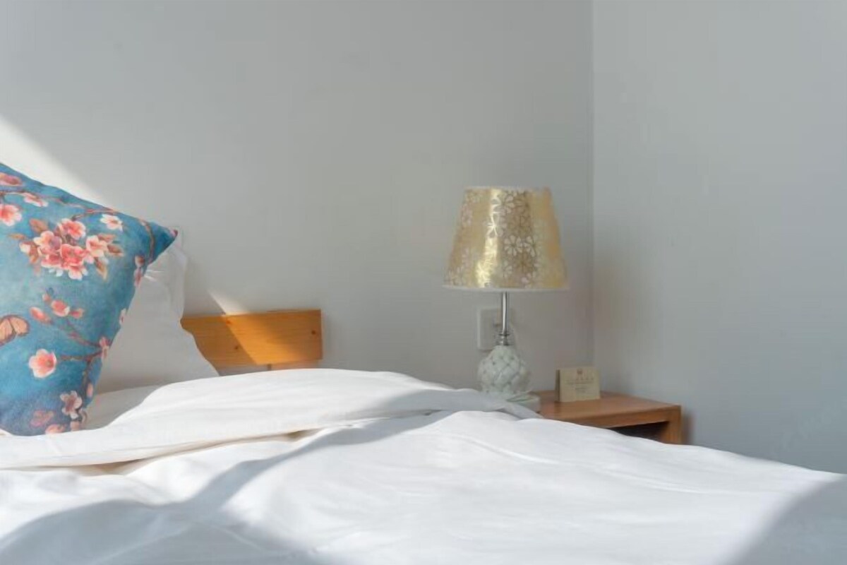 Una cama bien hecha con ropa blanca, una almohada con motivos florales azules y una mesita de noche de madera con una lámpara.
