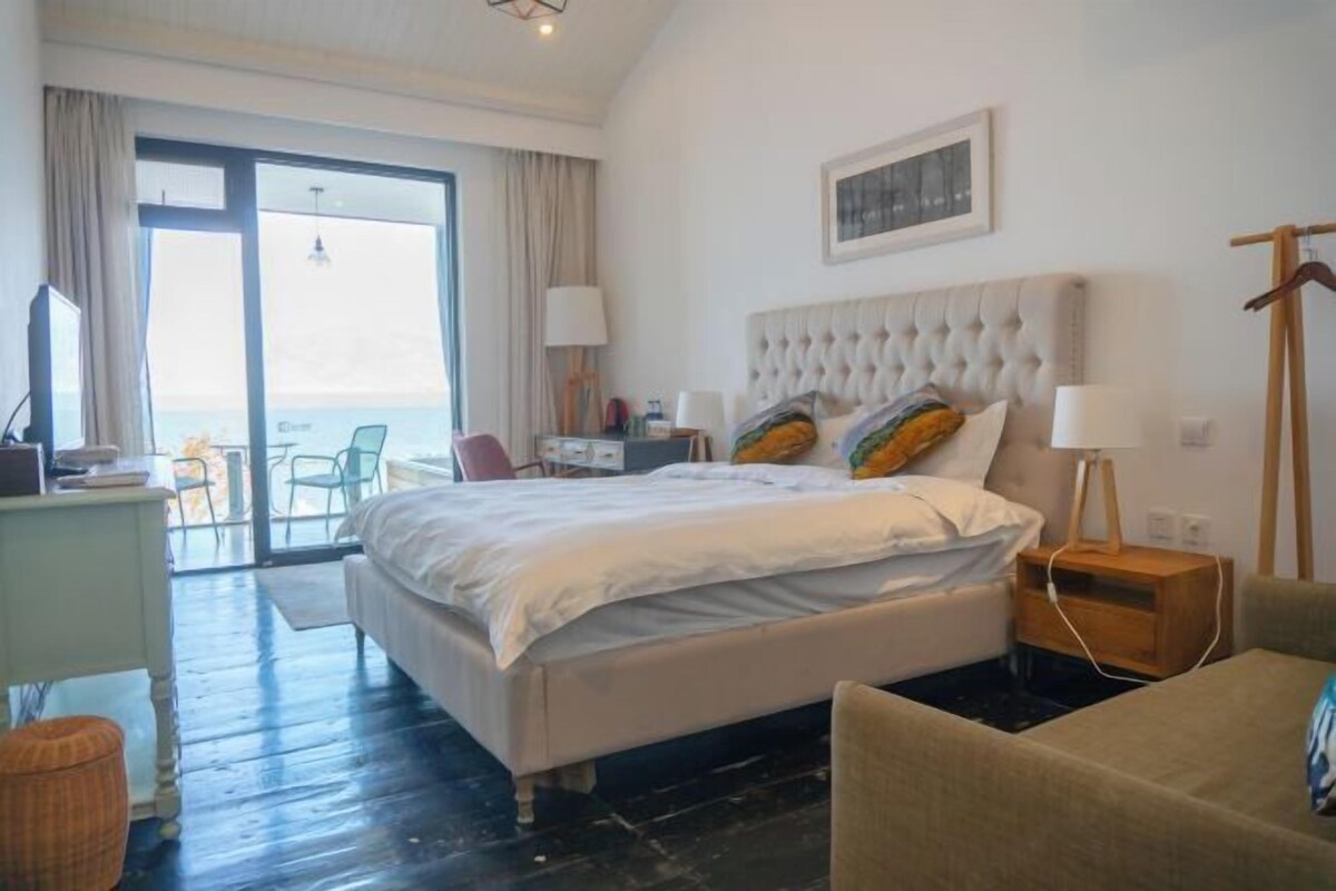 Habitación de hotel con una cama grande, un escritorio, una silla y vistas al mar a través de la puerta de cristal.