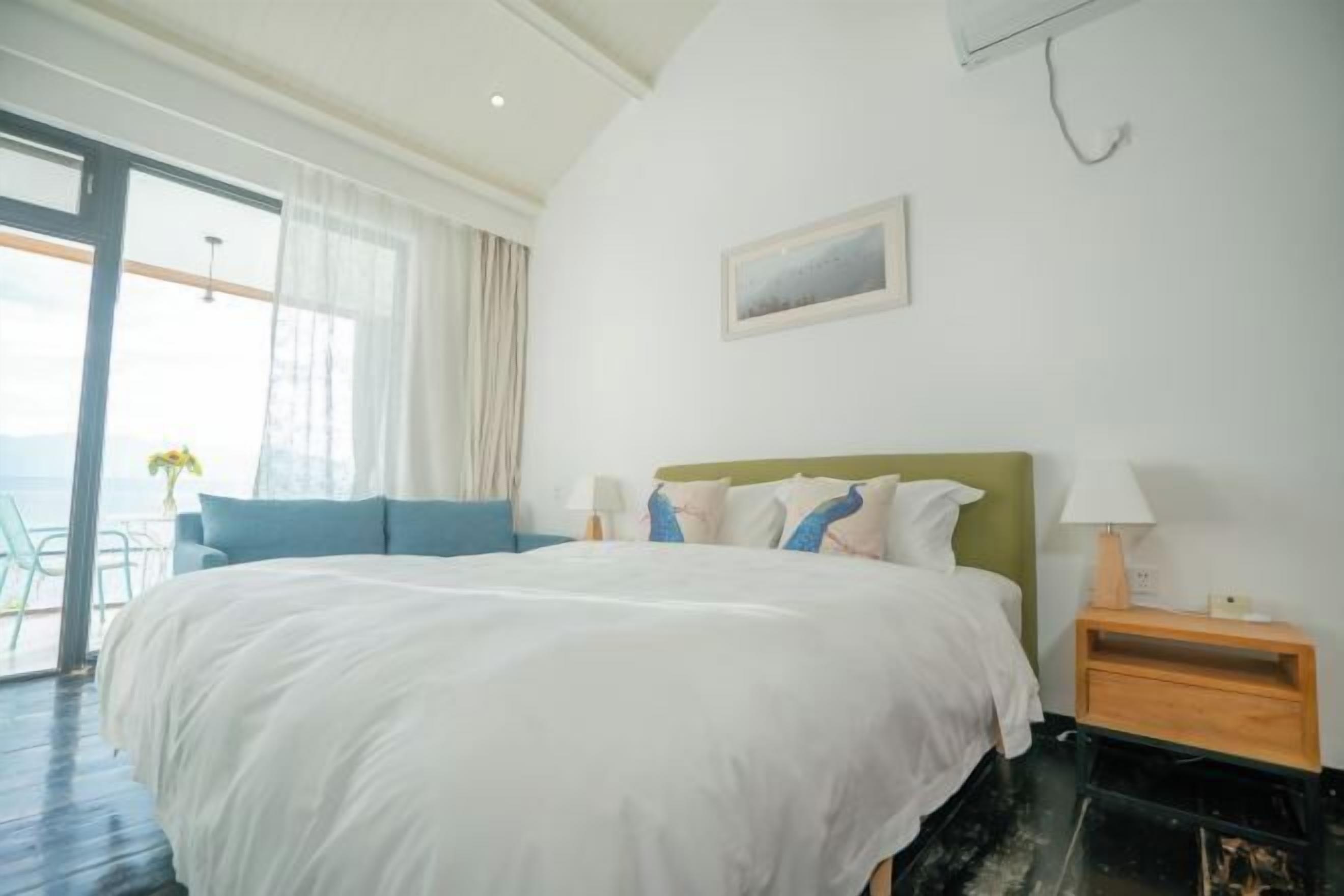 A bedroom with a large bed, a nightstand, a lamp, a painting on the wall, and a view through a window.
