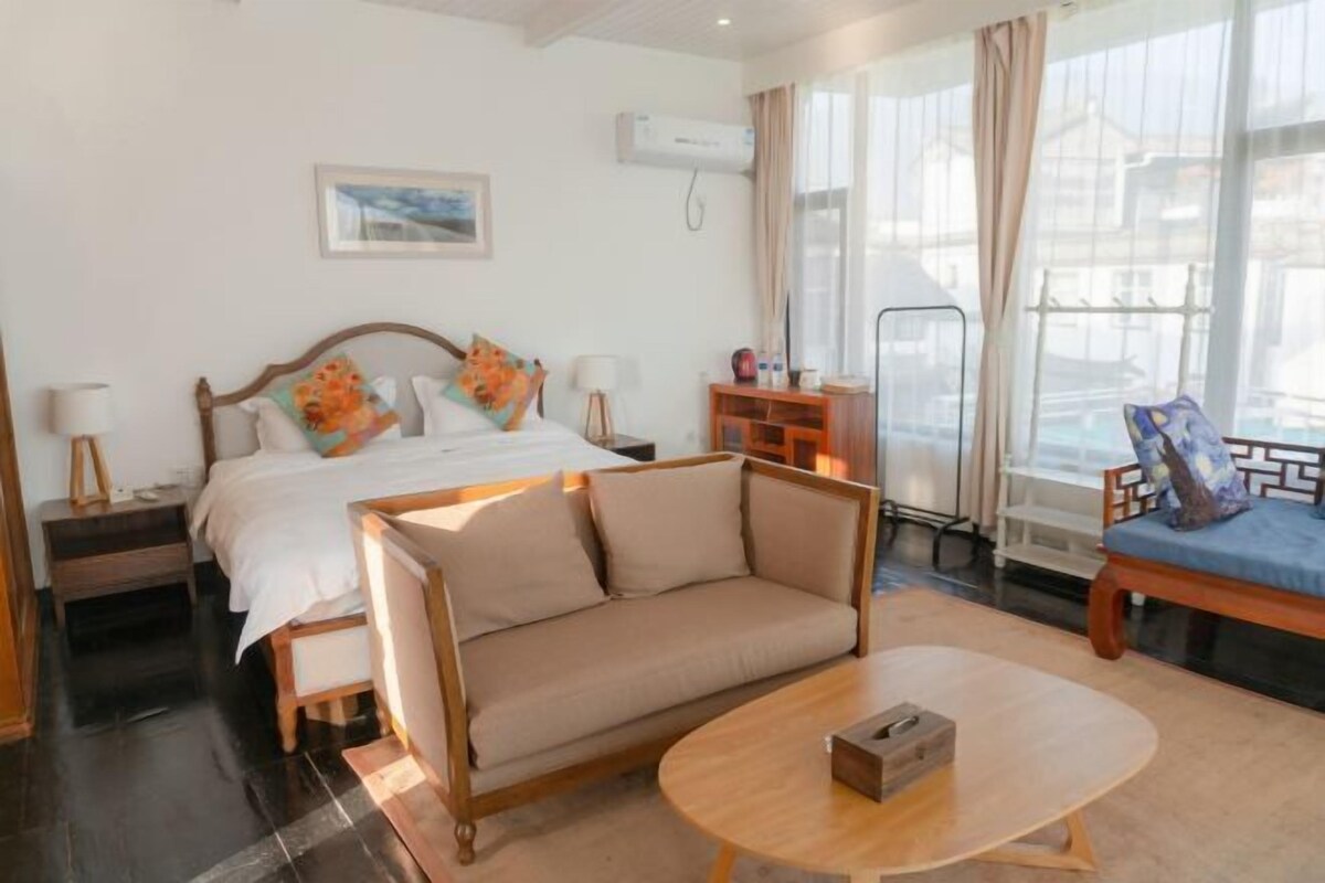 Habitación de hotel con cama, sofá, mesa de centro, mesita de noche, lámpara, televisor y ventana con cortinas.