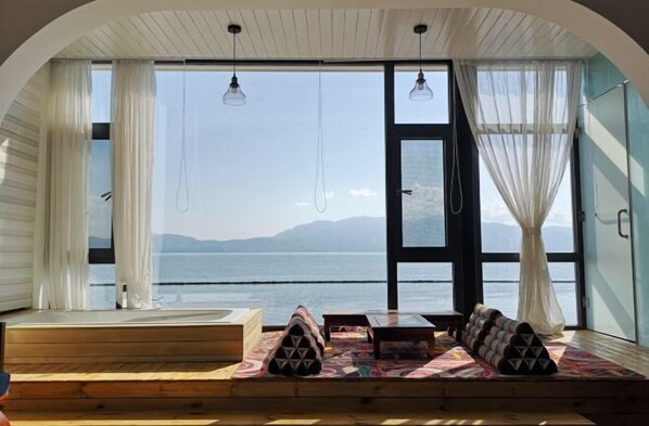 Habitación con un amplio ventanal que da a un cuerpo de agua y montañas, piso de madera y una zona de estar con cojines.