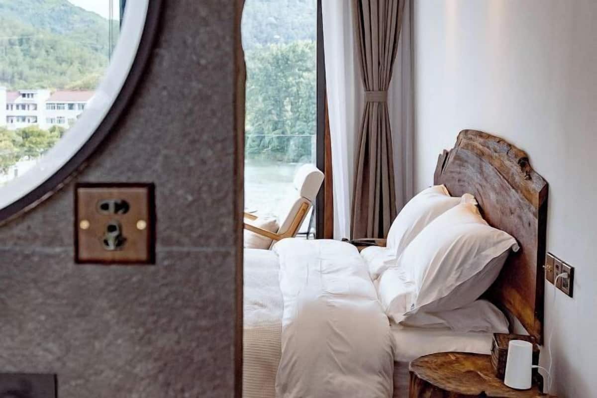 A bedroom with a wooden headboard, white bedding, a bedside table with a lamp, and a view of greenery outside.