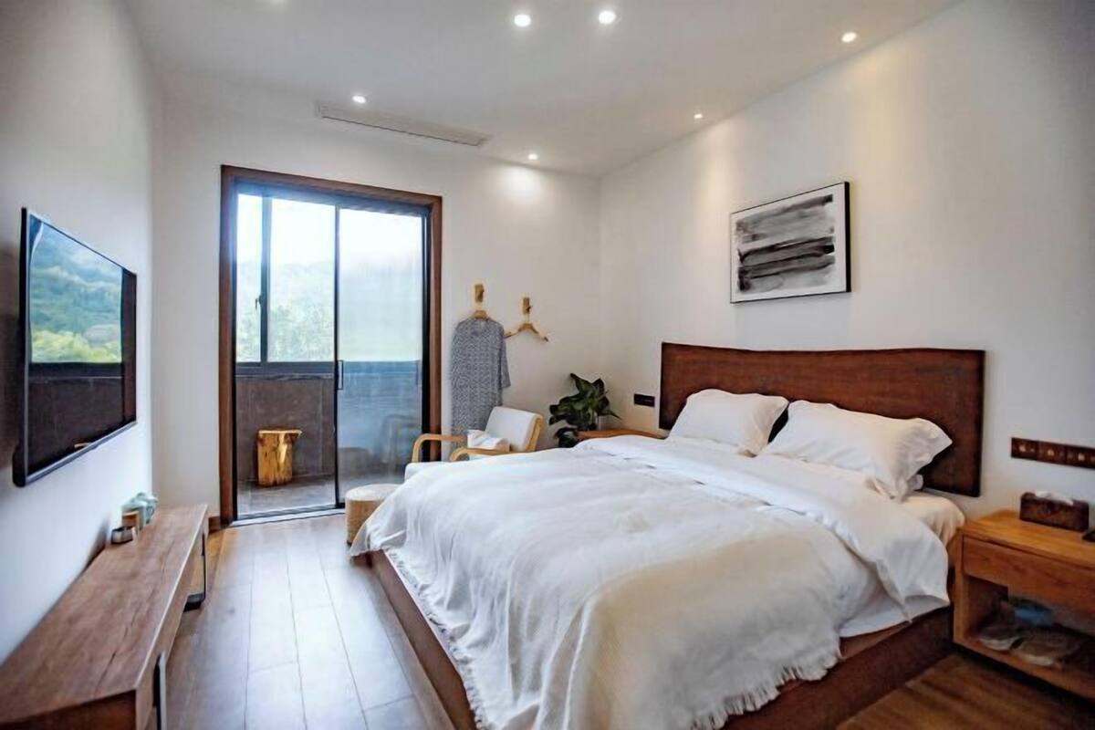 A bedroom with a large bed, a wooden headboard, a bedside table, a bench, a chair, and a view through a glass door.