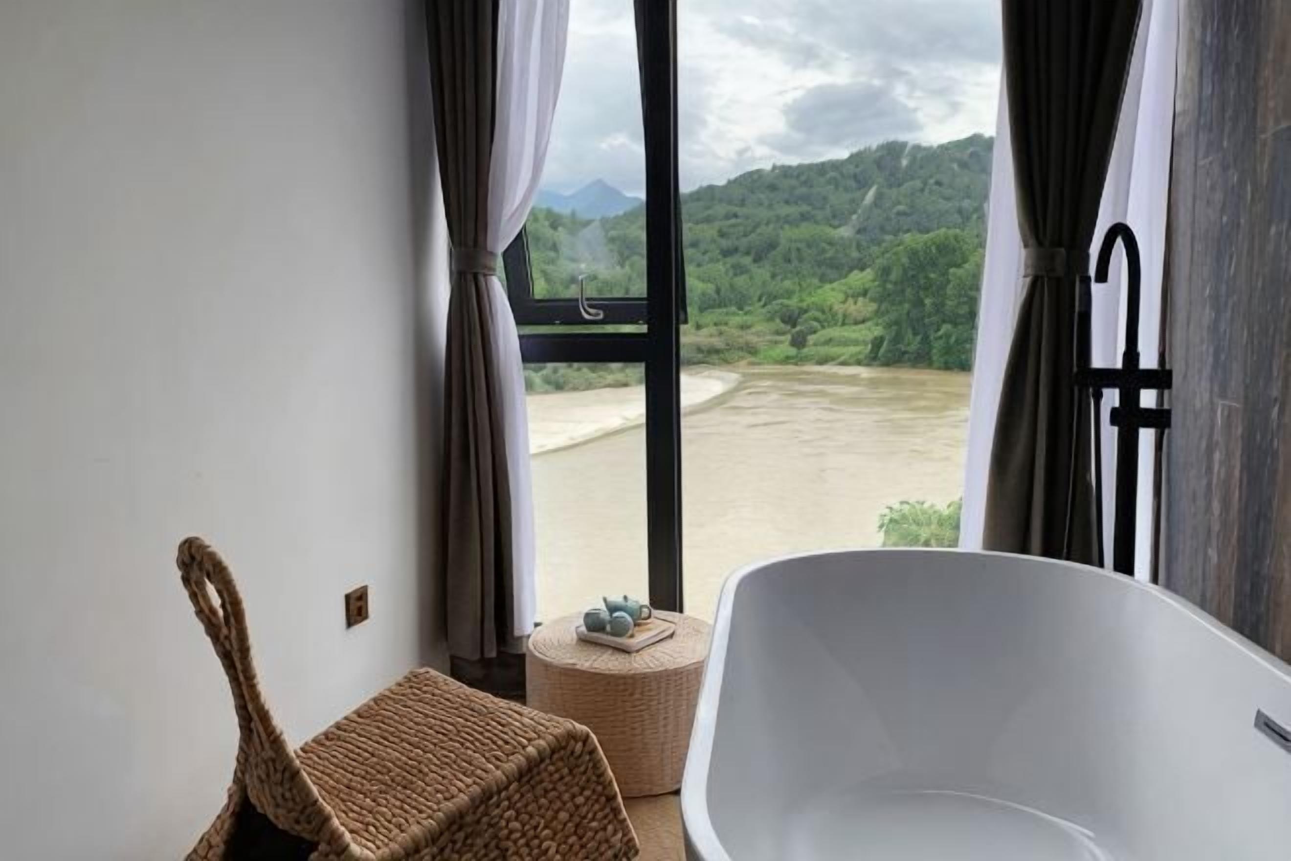 A room with a bathtub, a wicker chair, and a view of a river and mountains.