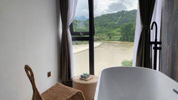 A room with a bathtub, a wicker chair, and a view of a river and mountains.