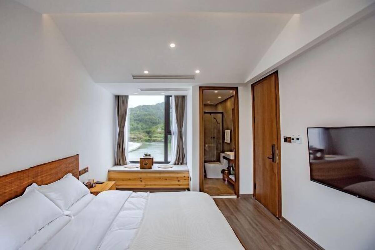 A bedroom with a large bed, a TV, a window with a view, and a wooden door leading to a bathroom.