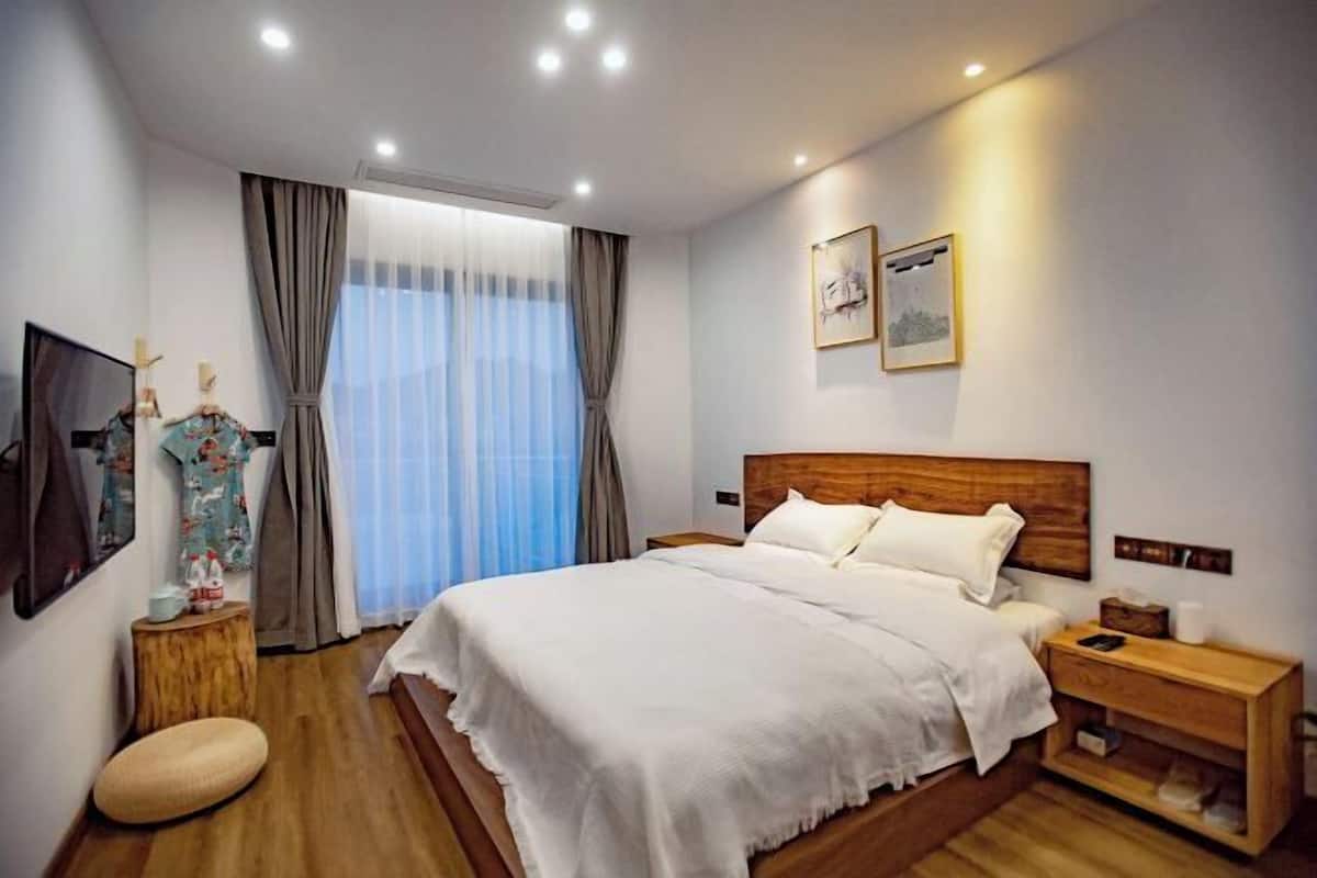 A hotel room with a large bed, wooden headboard, bedside tables, a nightstand, a wooden stool, and a large window with curtains.