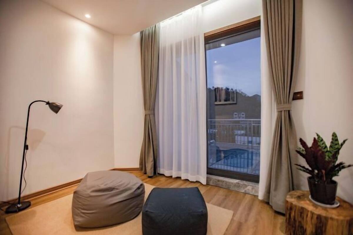 A room with a large window, a floor lamp, a bean bag, a small wooden table with a plant, and a view of a balcony.