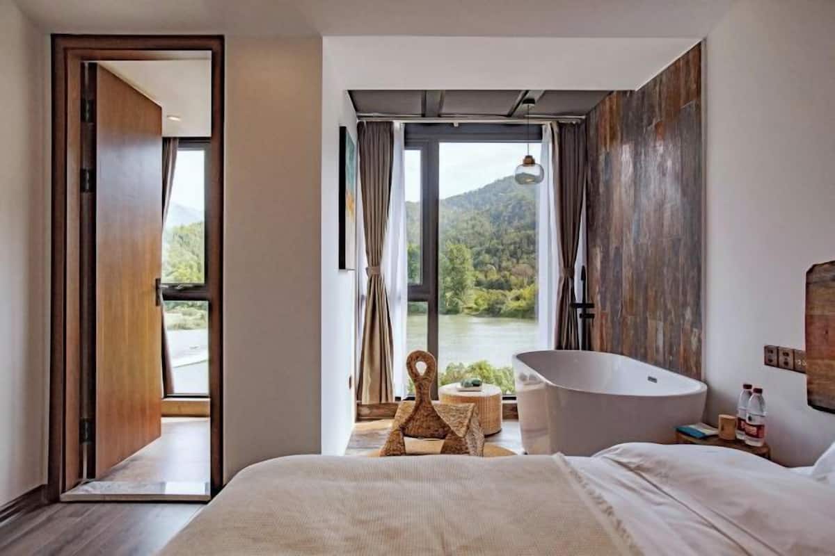 A modern bedroom with a large bed, a bathtub, and a view of a lake and mountains.