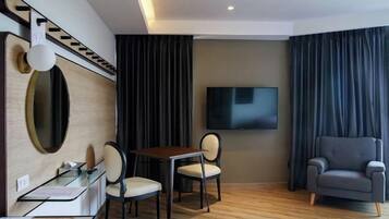 A hotel room with a wooden floor, a desk with a chair, a television mounted on the wall, and a sofa.