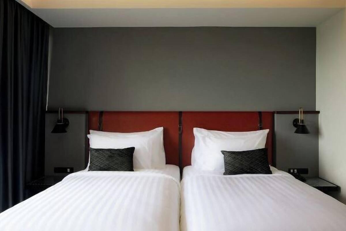 Un lit double avec du linge de lit blanc, deux tables de chevet avec des lampes, une tête de lit rouge et des rideaux foncés.