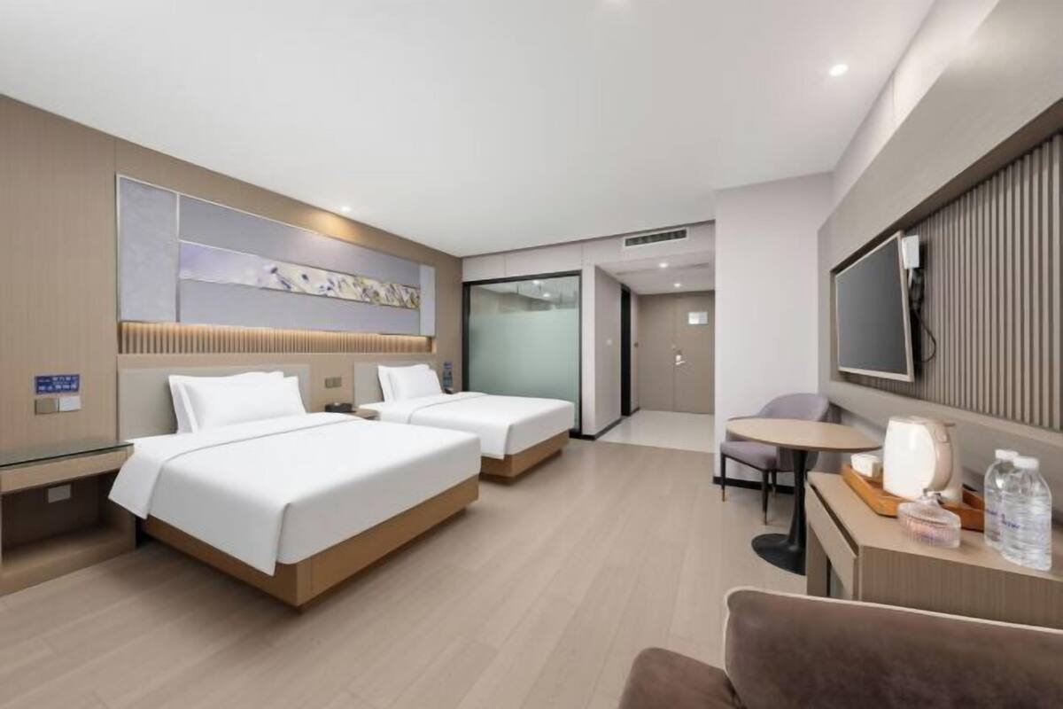 Ein Hotelzimmer mit zwei Betten, einem Schreibtisch, einem Stuhl, einem Fernseher und einer Glastrennwand.