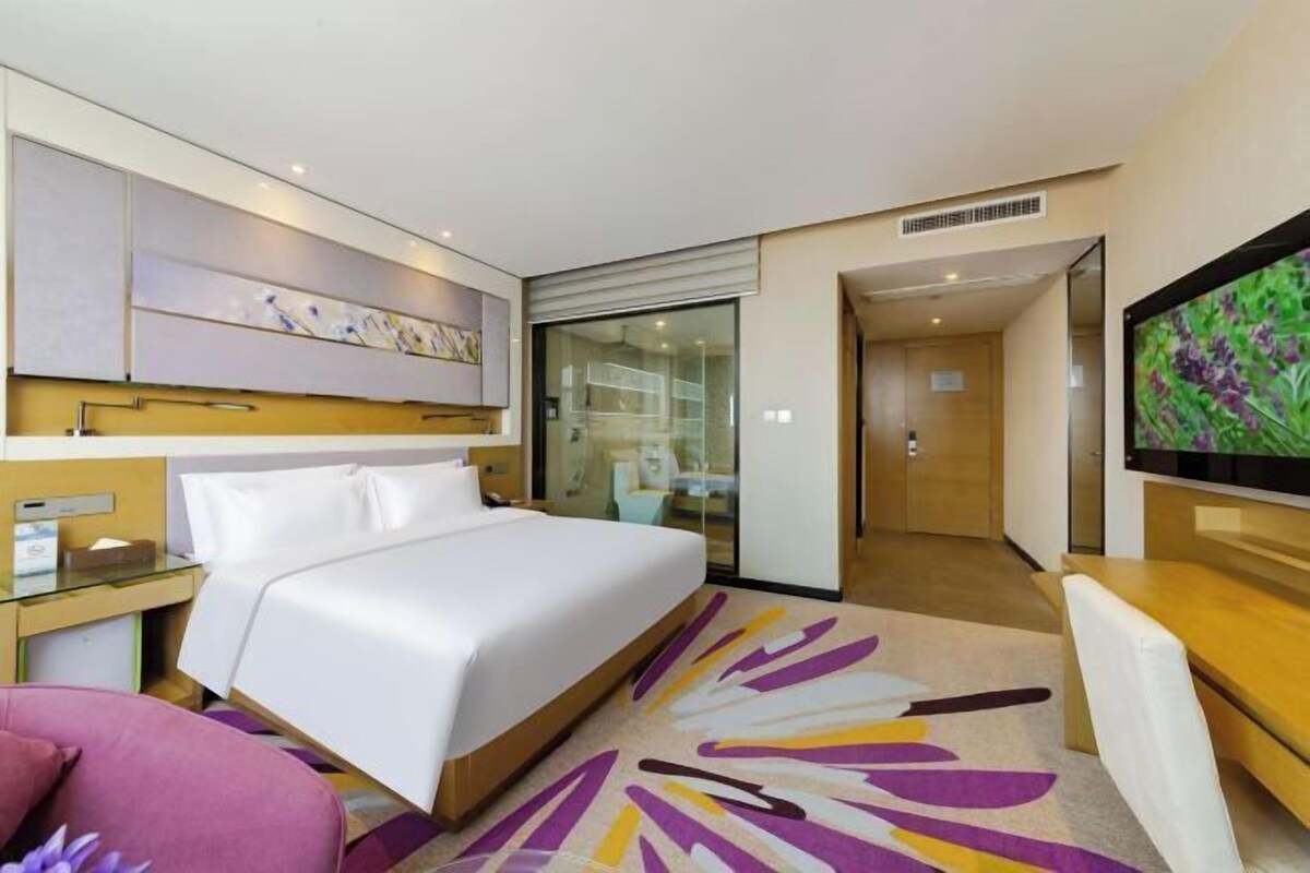 A hotel room with a bed, a desk with a computer, a TV, and a glass partition.
