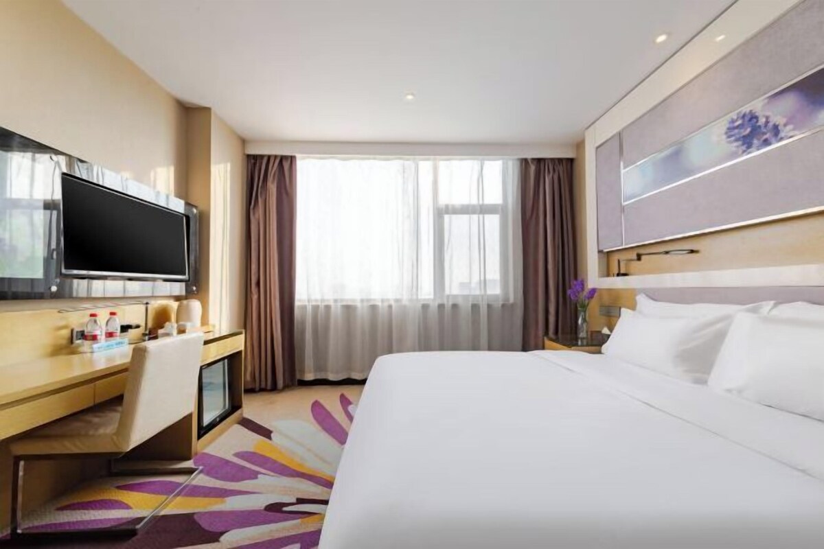 A hotel room with a bed, a desk with a TV, a window with curtains, and a painting on the wall.