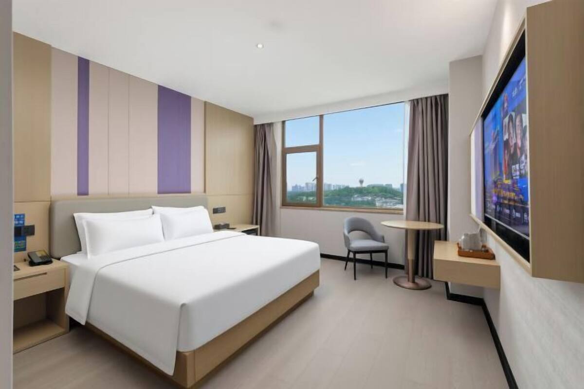 Ein Hotelzimmer mit Bett, Schreibtisch, Stuhl, Fernseher und einem Fenster mit Blick auf die Stadt.