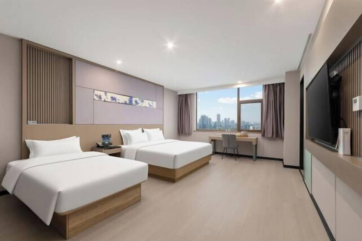 Ein Hotelzimmer mit zwei Betten, einem Schreibtisch, einem Sessel, einem Fernseher und Blick auf die Stadt.