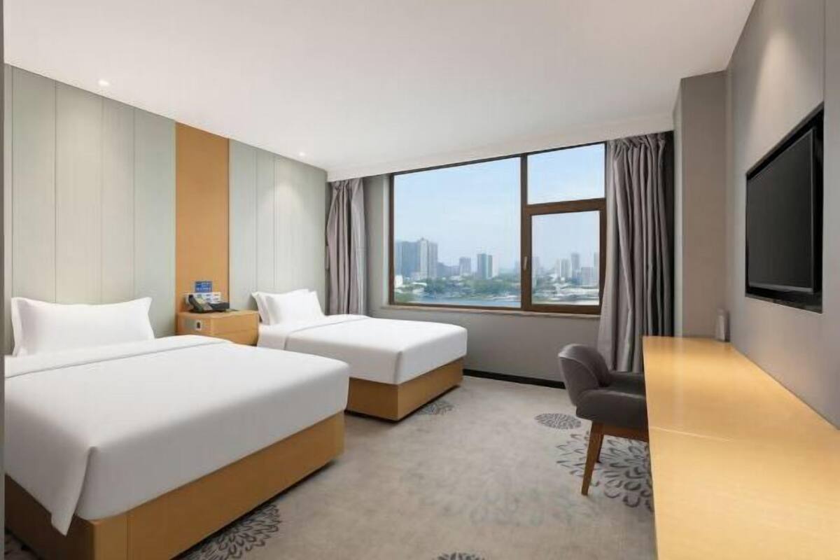 Ein Hotelzimmer mit zwei Betten, einem Schreibtisch, einem Sessel, einem Fernseher und einem großen Fenster mit Blick auf die Stadt.
