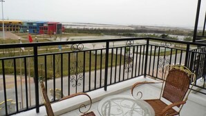 Un balcón con una mesa de vidrio y dos sillas de mimbre, con vistas a una zona costera con edificios y una bandera.