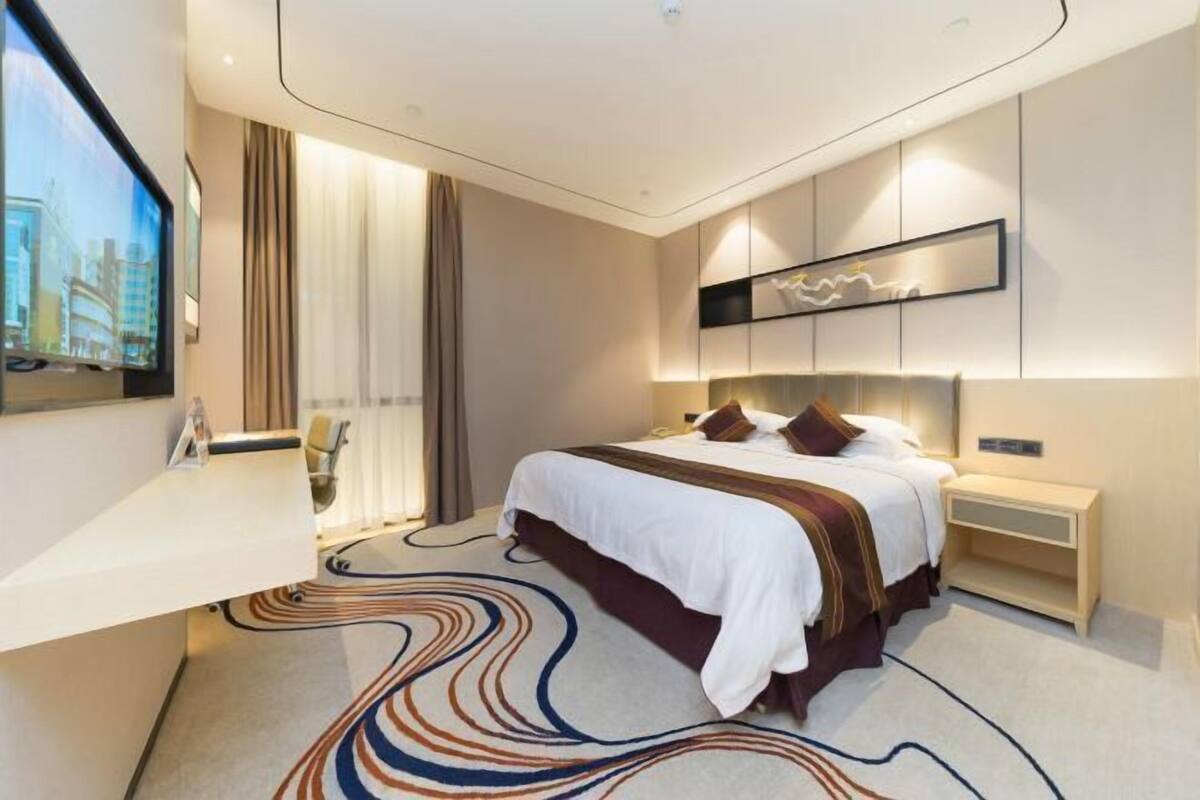 A modern hotel room with a large bed, a desk, a TV, and a view of the cityscape.