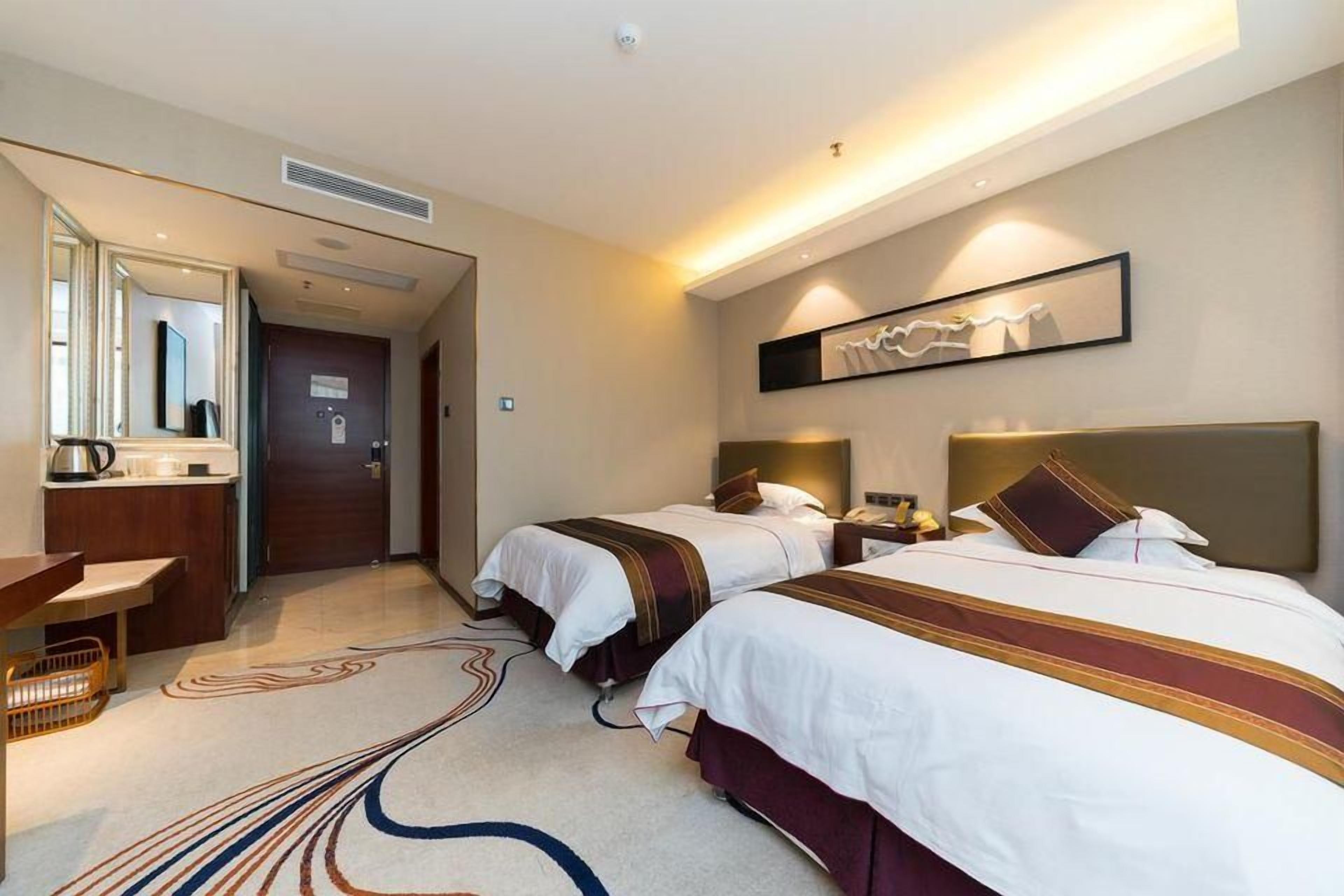 A hotel room with two beds, a desk, a mirror, and a wall-mounted artwork.