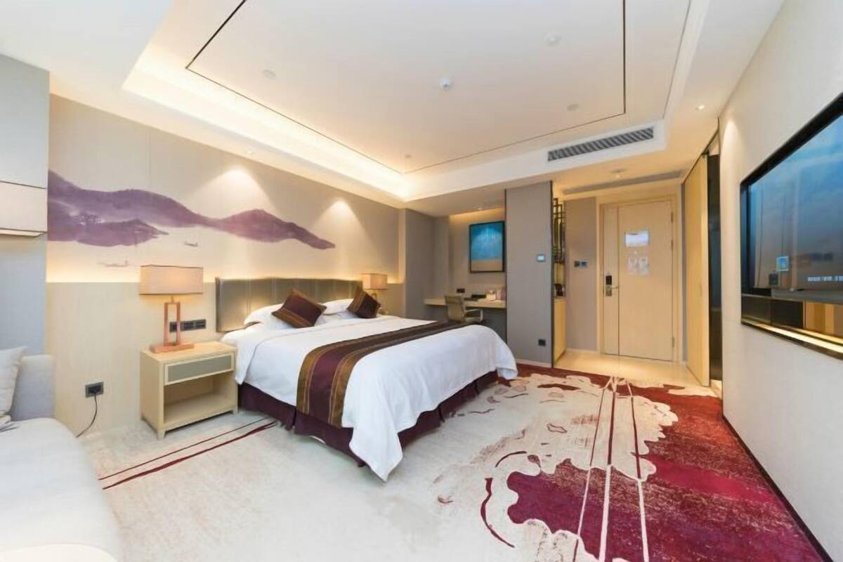 A modern hotel room with a large bed, bedside tables, a TV, and a carpeted floor.
