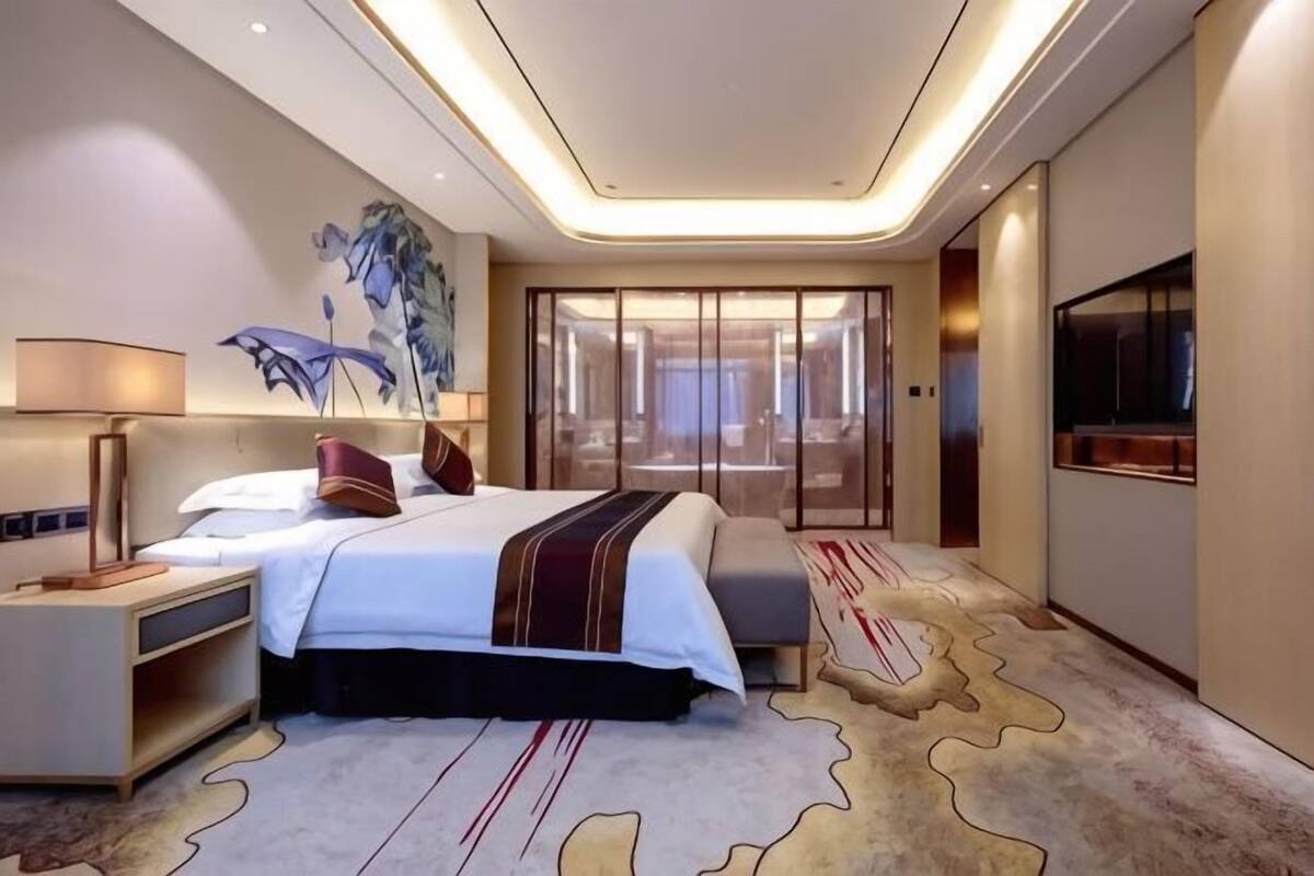 A modern hotel room with a large bed, a desk, a TV, and a view of the city through the window.