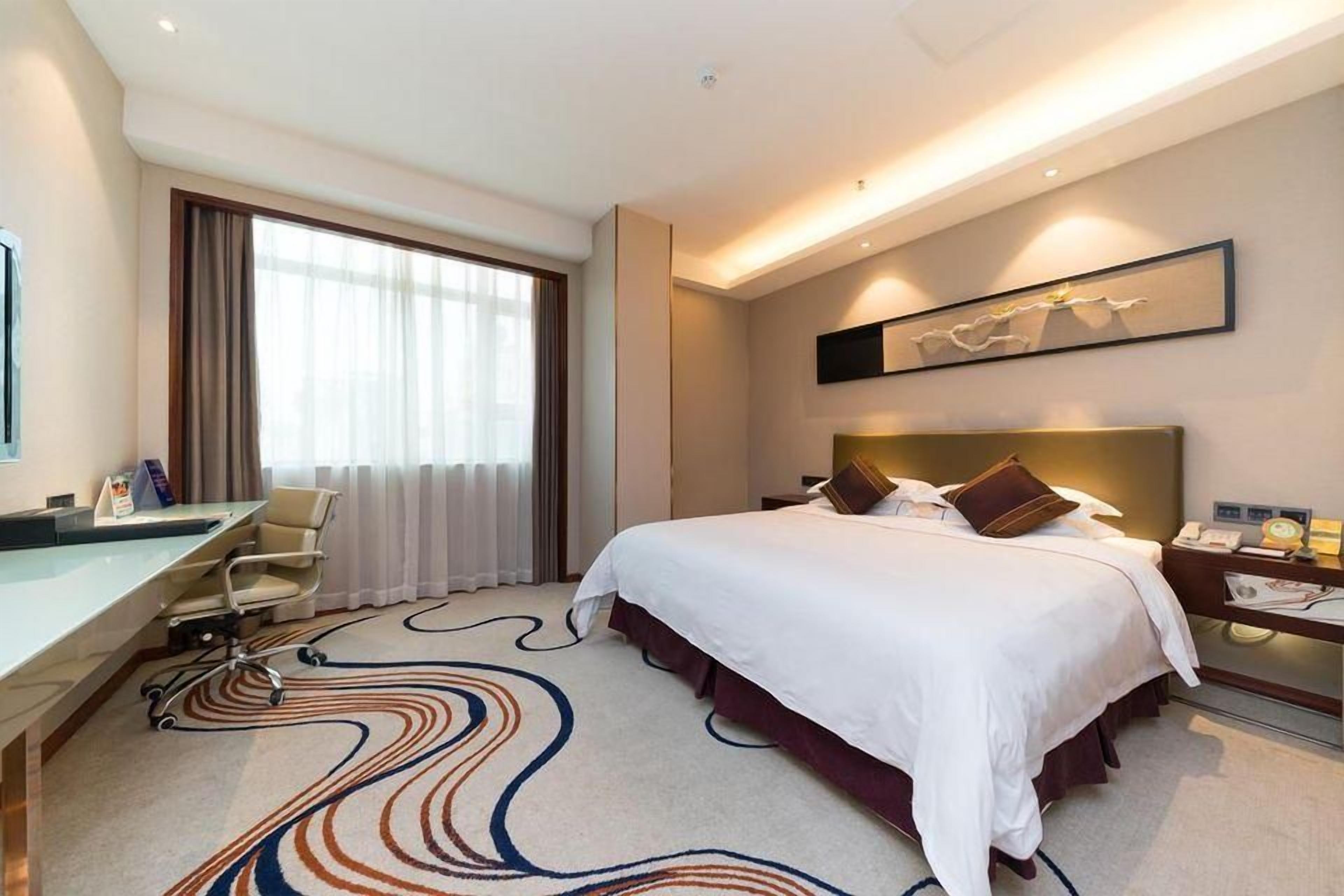 Habitación de hotel con cama, escritorio, silla y un ventanal con cortinas.