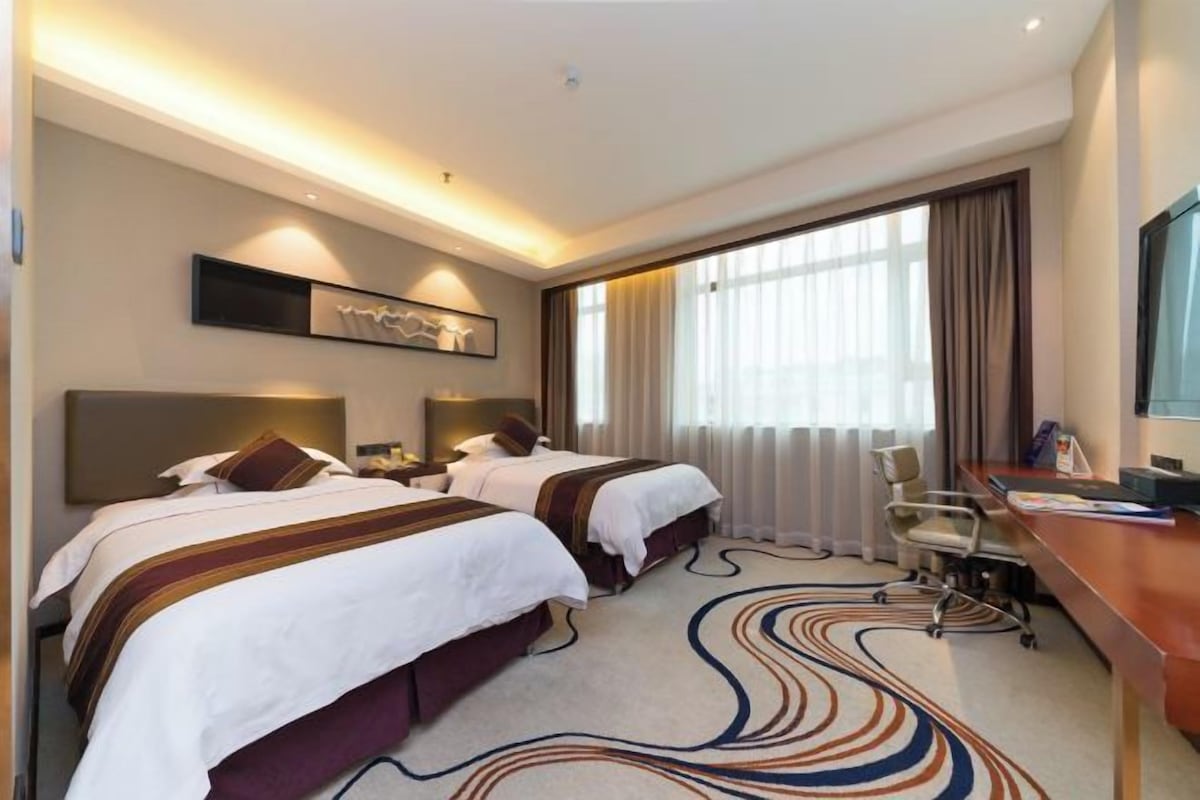 A hotel room with two beds, a desk, a chair, and a large window with curtains.