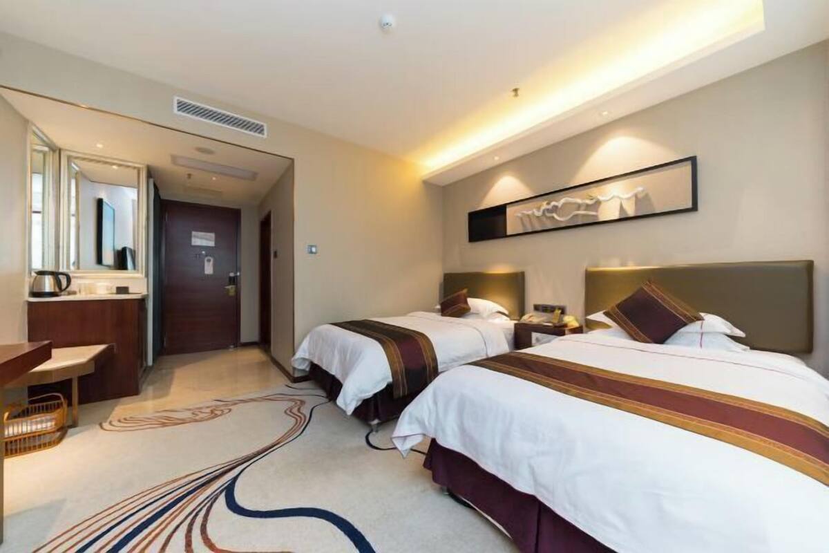 A hotel room with two beds, a desk, a mirror, and a wall-mounted artwork.