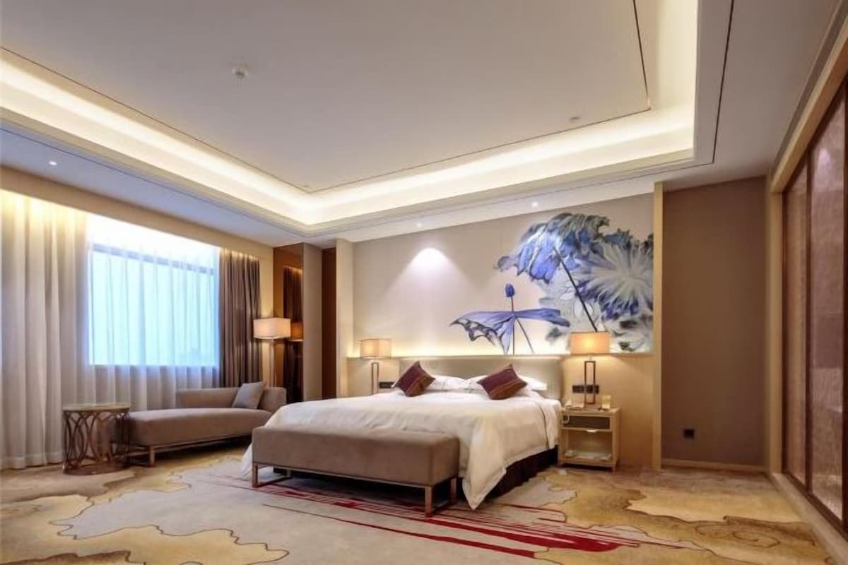 A hotel room with a bed, bedside tables, a sofa, a large window with curtains, and a wall-mounted artwork.