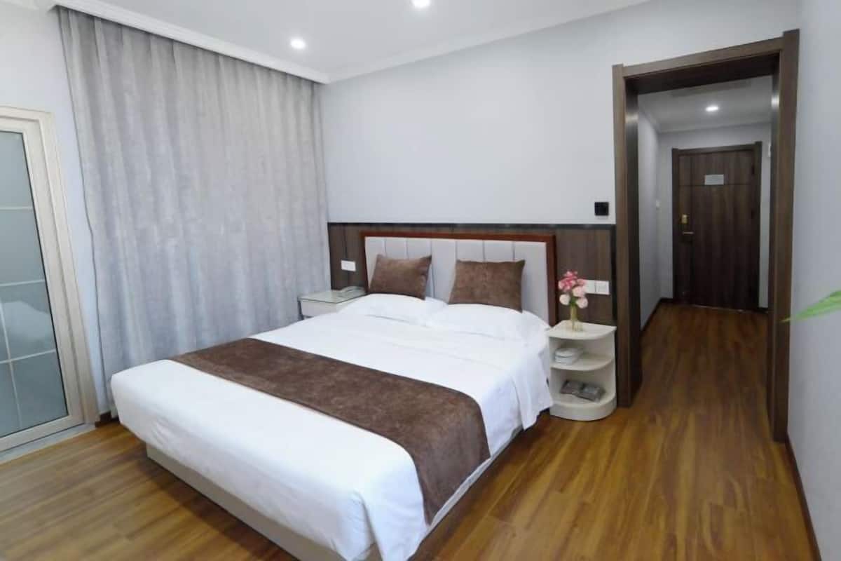 A hotel room with a large bed, a nightstand, a bedside table with a vase of flowers, and a wooden floor.