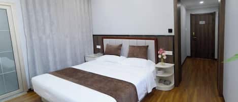 A hotel room with a large bed, a nightstand, a bedside table with a vase of flowers, and a wooden floor.