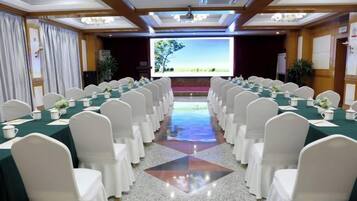 Une salle de conférence avec des rangées de chaises blanches, des tables vertes et un grand écran à l’avant.