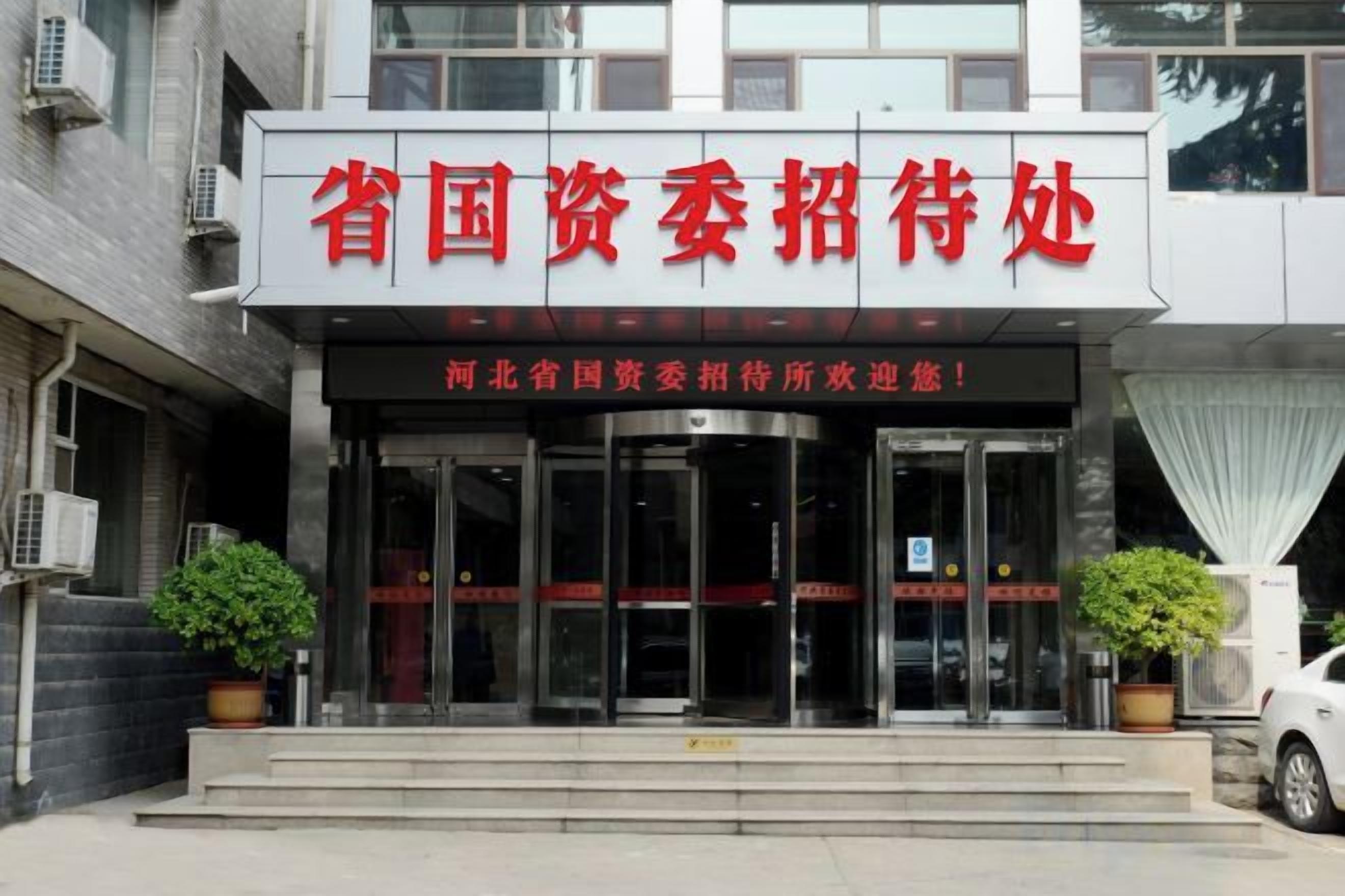 A building entrance with a sign in red and black, two potted plants, and a car parked outside.