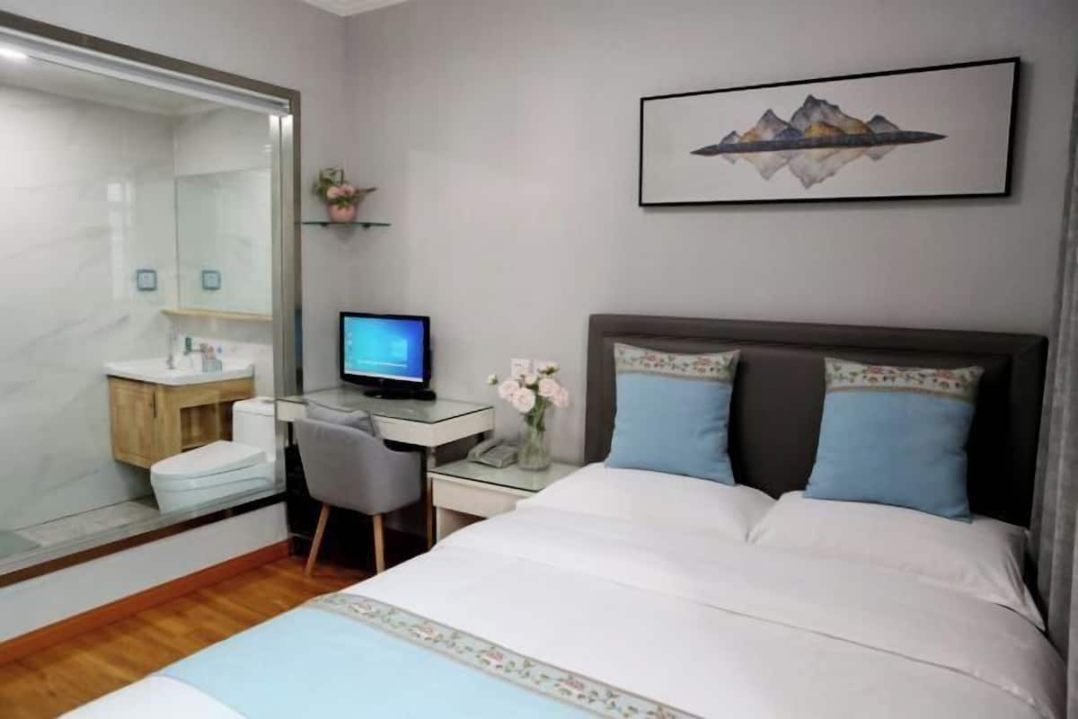 A hotel room with a bed, a desk with a computer, a mirror, a bathroom, and a framed picture on the wall.