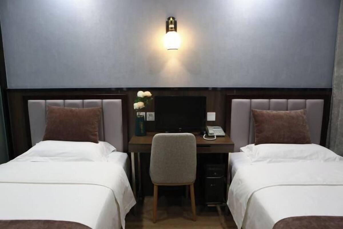 A hotel room with two beds, a desk with a computer, a chair, and a wall-mounted light.