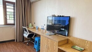 Une chambre d’hôtel avec un bureau en bois, une télévision, une chaise, un tabouret bleu et une boîte rouge.