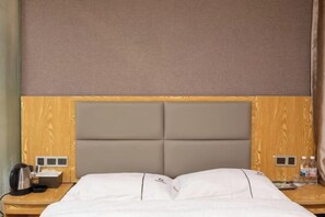 Quarto de hotel com cabeceira de madeira, cama com roupa de cama branca, mesa de cabeceira com telefone e chaleira, e uma pequena mesa de apoio com abajur e garrafa.