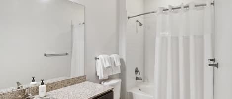 Un baño con lavamanos de encimera de granito, un espejo, inodoro, bañera y cortina de ducha.