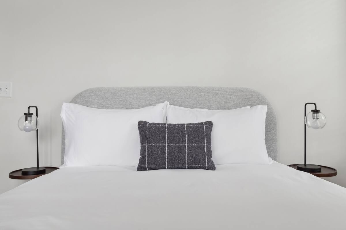 Ein ordentlich bezogenes Bett mit weißen Leinen, einem grauen Kopfteil und zwei Nachttischlampen.