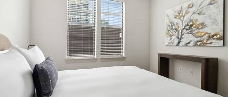 A bedroom with a bed, two windows with blinds, a painting on the wall, and a small wooden table.