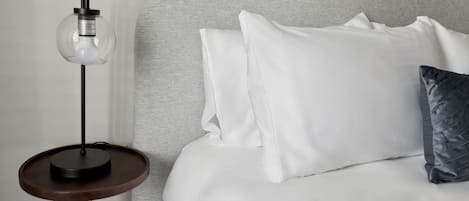 A bed with white bedding and a dark gray headboard, a small round table with a lamp, and a dark gray pillow.