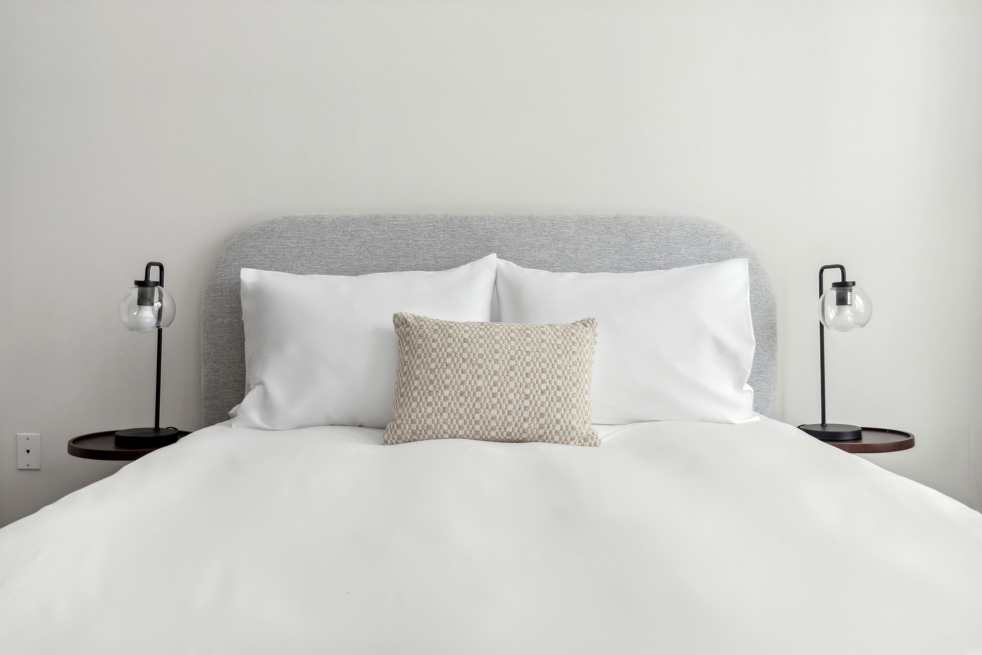 Een net opgemaakt bed met witte lakens, een grijze hoofdbord en twee bedlampjes.