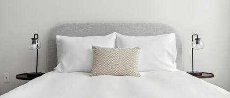 Een net opgemaakt bed met witte lakens, een grijze hoofdbord en twee bedlampjes.