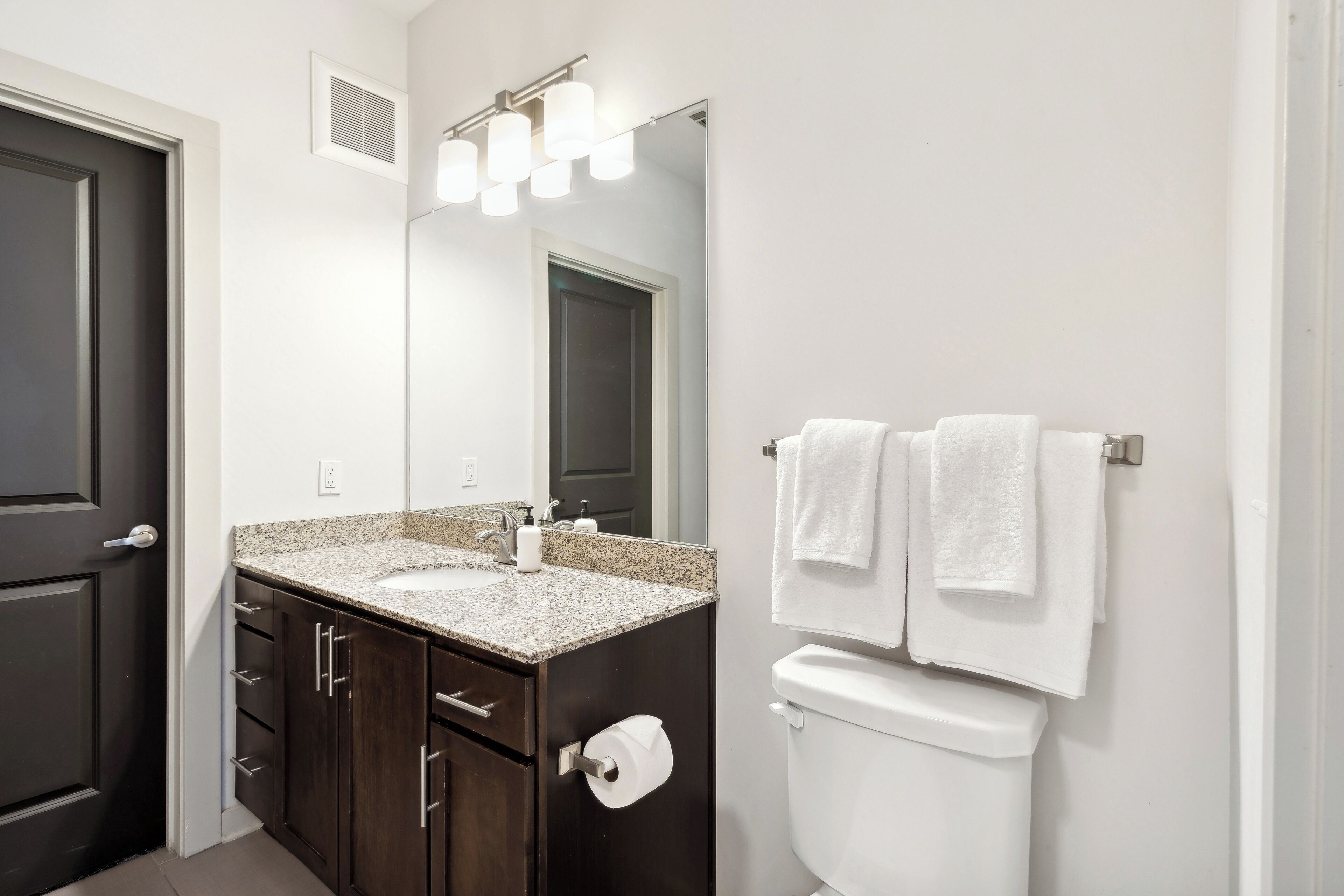 A modern bathroom with a dark wood vanity, a granite countertop, a white toilet, and a mirror with lights.