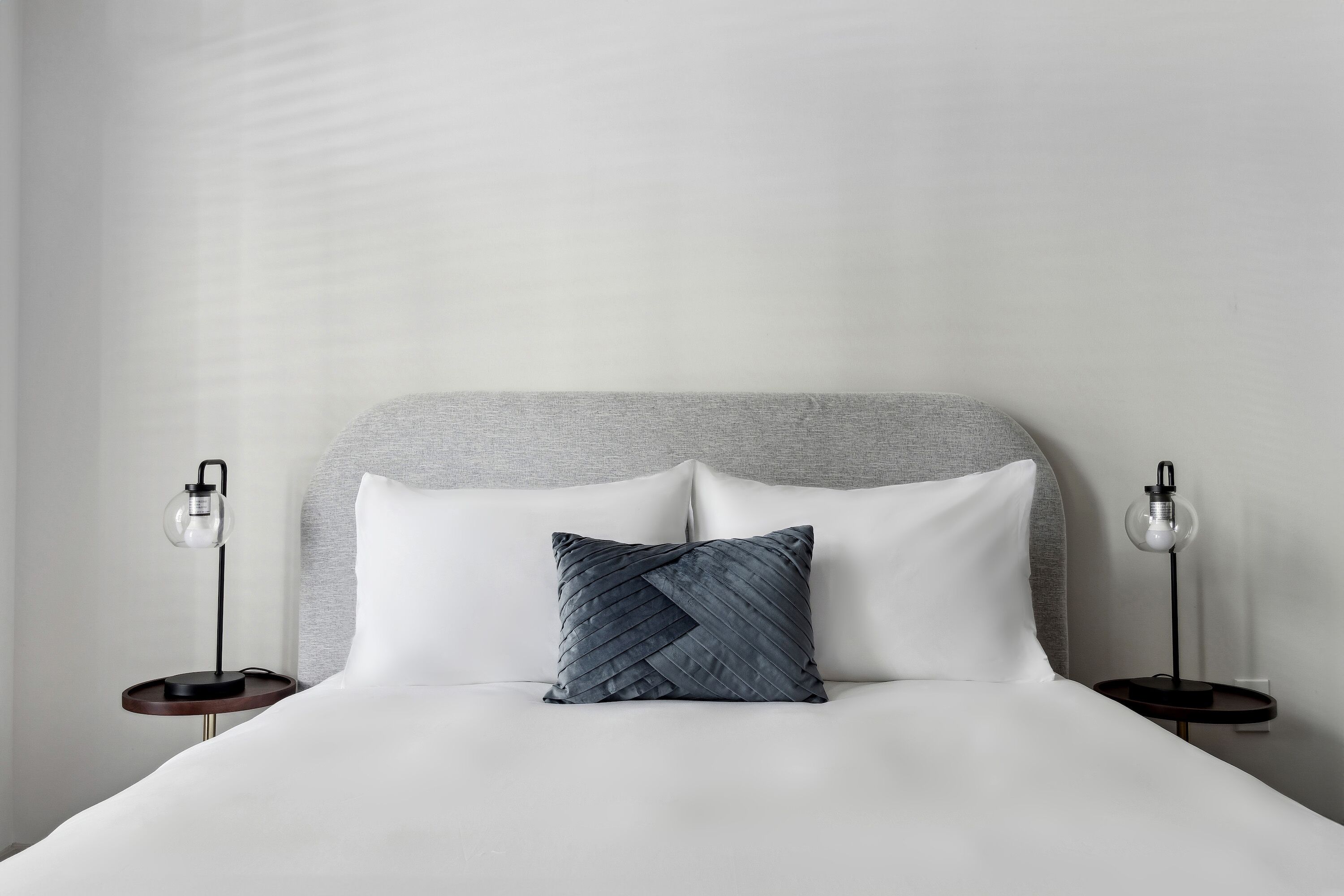 A neatly made bed with a grey headboard, two bedside tables with lamps, and a grey pillow.