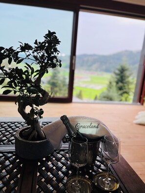 Une table ornée d’un bonsaï, d’une bouteille de champagne Graceland et de deux verres de vin, le tout placé devant un paysage montagneux pittoresque.