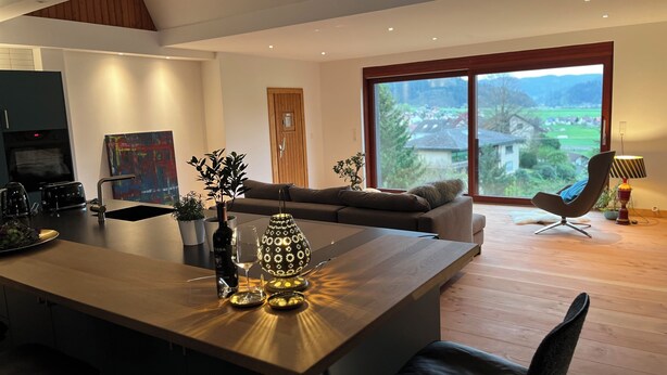 A modern kitchen with a central island, a dining area with a sofa, and a large window offering a view of the outdoors. - Graceland in the Black Forest (Fischerbach)