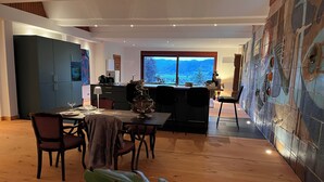 A modern dining area with a wooden table and chairs, a kitchen in the background, and a large window offering a view of the mountains. - Graceland in the Black Forest (Fischerbach)