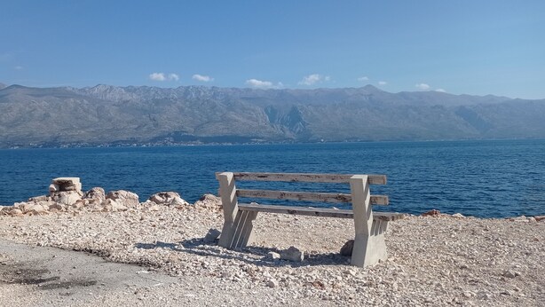 A bench on a rocky shore with a view of mountains and a body of water. - Apartment Tina (Ražanac)